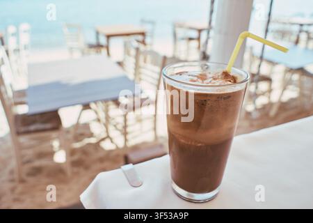Nahaufnahme eines erfrischenden Eiskaffees in einem hohen Glas mit einem gelben Strohhalm, der auf einem weißen Tisch in einem Strandcafé platziert ist, mit verschwommenen Tischen und Stühlen Stockfoto