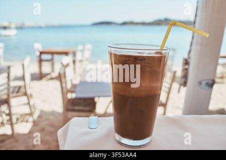 Eine Nahaufnahme eines erfrischenden Eiskaffees, serviert in einem hohen Glas mit einem gelben Strohhalm, der auf einem Tisch in der Nähe des Meeres und eines Sandstrands in G steht Stockfoto