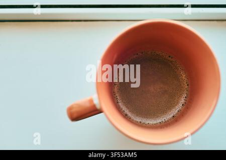 Eine Nahaufnahme aus hohem Winkel, die direkt in eine orangefarbene Keramiktasse blickt, die mit frisch gebrühtem heißem Kaffee gefüllt ist und eine leichte Schicht Schaum aufweist Stockfoto