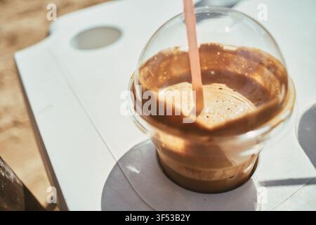 Eine Nahaufnahme eines kalten Eiskaffees in einem klaren Plastikbecher zum Mitnehmen mit einem rosa Strohhalm, der auf einem weißen Tisch unter hellem Sonnenlicht steht, Gre Stockfoto