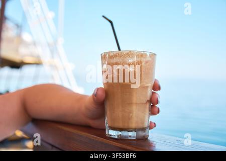 Nahaufnahme einer Person, die ein erfrischendes Glas traditionellen Eiskaffee-Frappe mit einem Strohhalm in der Nähe des Meeres in Griechenland hält Stockfoto