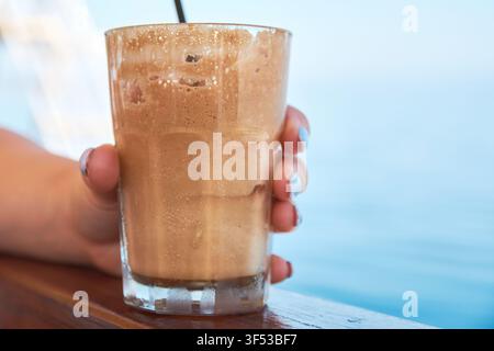 Nahaufnahme einer Person, die ein hohes Glas mit einem kalten Eiskaffee-Frappe mit dickem Schaum hält, vor einem hellblauen Meereshintergrund in Gree Stockfoto