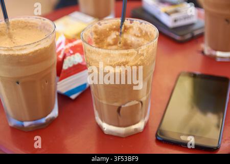 Nahaufnahme von erfrischenden Eiskaffee-Getränken in hohen Gläsern, die auf einem roten Café-Tisch neben einem Smartphone und Zuckerpäckchen während eines rel platziert sind Stockfoto