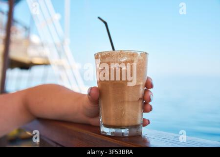 Nahaufnahme einer Hand, die ein hohes Glas mit kaltem, schaumigem Kaffeegetränk mit einem schwarzen Strohhalm vor einem hellen Meereshintergrund in Griechenland hält. Stockfoto