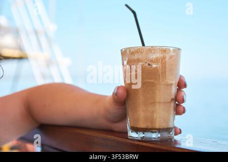 Nahaufnahme einer Hand, die ein hohes Glas mit Eiskaffee-Frappe und einem Strohhalm hält, das auf einem Holzgeländer platziert ist, mit einem unscharfen blauen Meereshintergrund, Griechenland Stockfoto