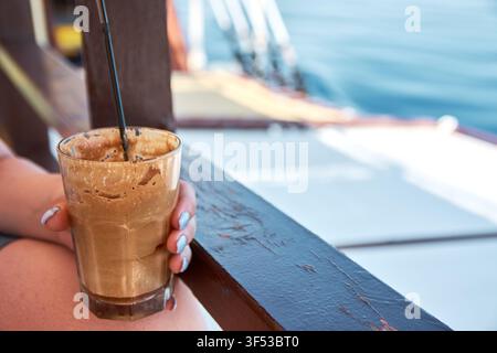 Nahaufnahme einer Person, die ein Glas mit einem eisigen, schaumigen Kaffee-Frappe mit einem Strohhalm hält, sitzend an einem Holzgeländer mit einem verschwommenen blauen Meereshintergrund in Stockfoto