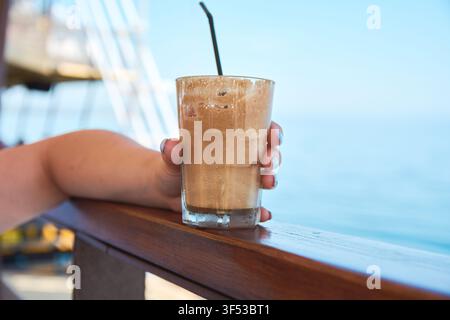 Nahaufnahme eines erfrischenden kalten Eiskaffee-Frappe-Getränks mit Schaum in einem Glas, das von einer Person auf einer Seeterrasse mit Meereshintergrund gehalten wird. Stockfoto
