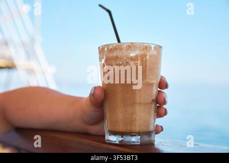 Nahaufnahme eines erfrischenden Eiskaffee-Frappé in der Hand mit Meereshintergrund in Griechenland, mit schaumiger Textur und einem Strohhalm. Stockfoto