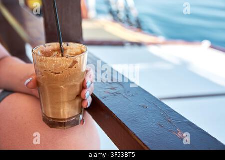 Nahaufnahme einer Person, die ein hohes Glas mit schaumigem Eiskaffee-Frappe mit einem schwarzen Strohhalm hält, während sie auf einem hölzernen Bootsdeck mit Meeresblick-Hintergrund sitzt Stockfoto