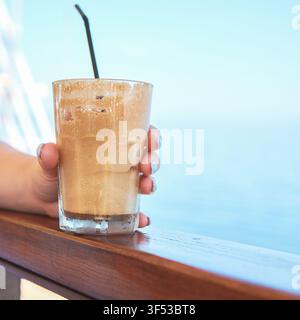 Nahaufnahme einer Person, die ein erfrischendes Glas kalten, schaumigen Eiskaffee-Frappe mit einem Strohhalm auf einem Holzgeländer mit Blick auf das Meer in Griechenland hält Stockfoto