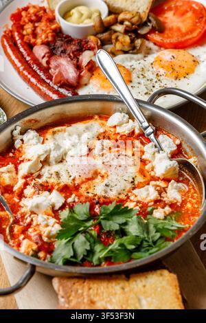 Traditionelle nahöstliche Shakshuka mit Fetakäse, serviert mit englischem Frühstück und Kaffee auf einem Holztisch Stockfoto