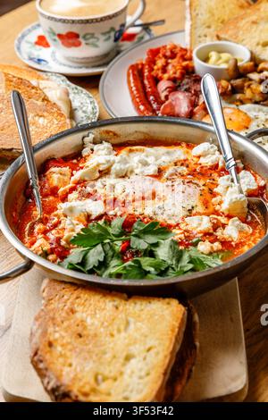Traditionelle nahöstliche Shakshuka mit Fetakäse, serviert mit englischem Frühstück und Kaffee auf einem Holztisch Stockfoto