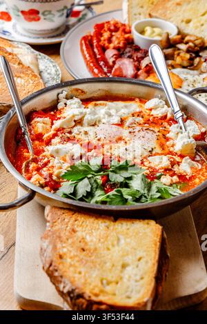 Traditionelle nahöstliche Shakshuka mit Fetakäse, serviert mit englischem Frühstück und Kaffee auf einem Holztisch Stockfoto