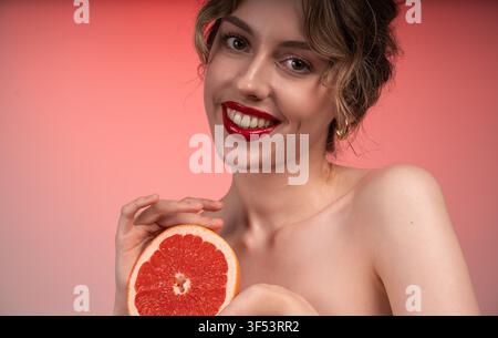 Junge Frau lächelt, hält Grapefruit, wirbt für gesunde Haut und natürliche Schönheit mit roten Lippen Stockfoto