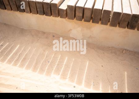 Nahaufnahme von verwitterten Holzlatten, die die Kante einer rustikalen Bank bilden, über windgepeitschtem hellem Sand und einer sonnenbeschienenen Verputzwand, warme Beige- und Brauntöne, s Stockfoto