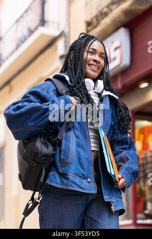 Junge afroamerikanische Studentin lächelt beim Spaziergang in einer Stadtstraße, trägt Rucksack und Bücher und genießt den urbanen Lebensstil Stockfoto