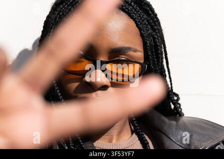Junge Frau, die mit der Hand ein Stoppschild mit Gesten hält, mit Sonnenbrille und Zöpfen Stockfoto