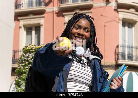 Glückliche Universitätsstudentin, die Bücher hält und einen grünen Apfel anbietet, um gesunde Ernährungsgewohnheiten für ein produktives akademisches Leben zu fördern Stockfoto