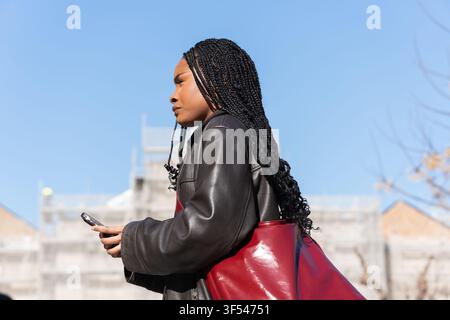 Junge schwarze Frau mit Zöpfen, die in der Stadt spazieren geht und ihr Smartphone benutzt Stockfoto