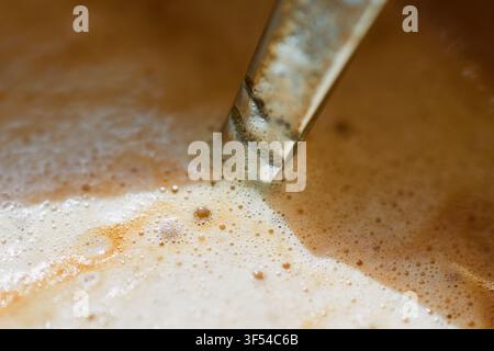 Extreme Makro-Nahaufnahme von reichem Kaffeeschaum mit winzigen Bläschen und einem Metalllöffel, der während der Zubereitung im heißen Getränk ruht Stockfoto