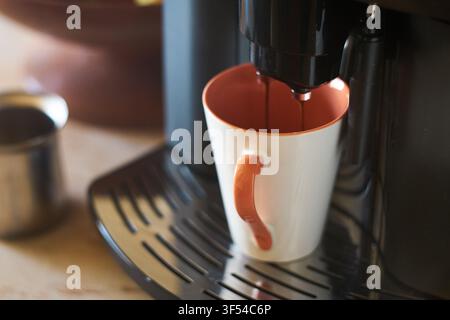 Nahaufnahme von reichem dunklem Espresso, der aus dem Auslauf einer modernen Kaffeemaschine in eine Keramiktasse fließt, die auf einem Tropftablett steht. Stockfoto