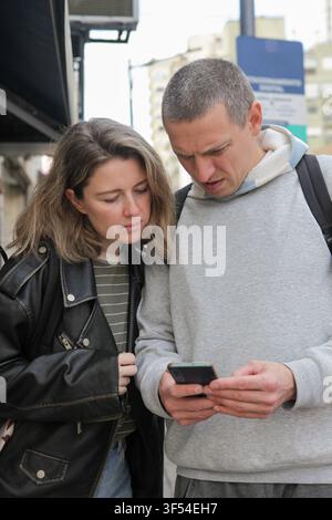 Mitte-alterliches Paar überprüft Informationen auf einem Smartphone zusammen, während es eine Straße in der Stadt entlanggeht. Stockfoto