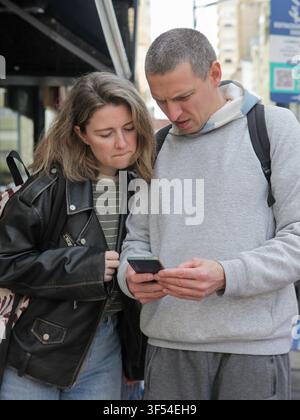 Mitte-alterliches Paar überprüft Informationen auf einem Smartphone zusammen, während es eine Straße in der Stadt entlanggeht. Stockfoto