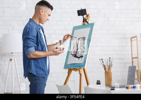 Lerne zu Hause während der Quarantäne Bilder zu zeichnen und online Unterricht zu erhalten. Junger Mann in Denimhemd zeichnet abstrakte Formen auf Leinwand und schaut sich ein Video an Stockfoto