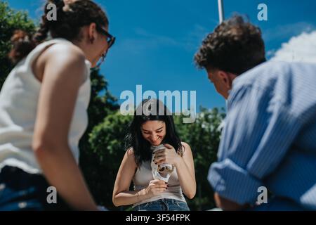 Freunde lachen im Freien, während eine junge Frau an einem sonnigen Tag einen schmelzenden Eisbecher hält Stockfoto