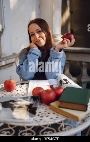 Lächelnde junge Frau sitzt an einem Tisch mit roten Äpfeln und Büchern, trägt einen gemütlichen blauen Strickpullover in einer entspannten Außencafé-Atmosphäre Stockfoto