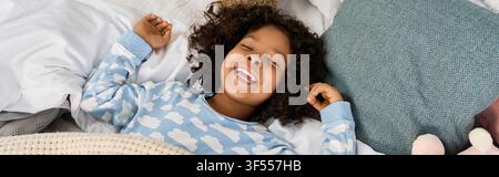 Ein süßer afroamerikanischer Junge lächelt in seinem kuscheligen Bett am Morgen, Banner Stockfoto