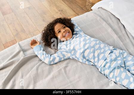 Ein fröhlicher afroamerikanischer Junge in süßen Pyjamas liegt auf seinem gemütlichen Bett Stockfoto