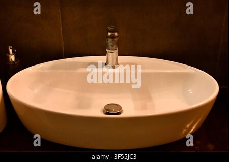 Nahaufnahme der Vorderseite eines modernen Keramik-Waschtischs mit einem Chrom-Wasserhahn, der auf einer dunklen Arbeitsplatte in einem stilvollen Badezimmer montiert ist Stockfoto