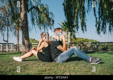 Paar sitzt Rücken an Rücken auf dem Gras, ignoriert sich gegenseitig und nutzt Smartphones Stockfoto