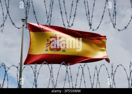 Spanische Flagge, die an einer Flaggenstange weht, gesehen hinter scharfem Stacheldraht vor einem bewölkten Himmel. Konzeptbild, das die europäische Einwanderungspolitik darstellt Stockfoto