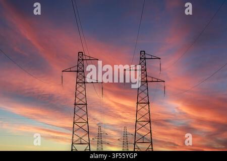 Niedrigwinkelaufnahme von Hochspannungsmasten-Silhouetten und Kabeln vor einem dramatischen Sonnenuntergangshimmel mit orangefarbenen und rosa Wolken. Stockfoto