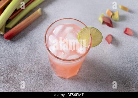Hausgemachte Rhabarberlimonade mit Eiswürfeln und frischer Limette, serviert in einem hohen Glas mit frischen Rhabarberstangen auf hellem Steinboden, erfrischender Sommerdrink Stockfoto
