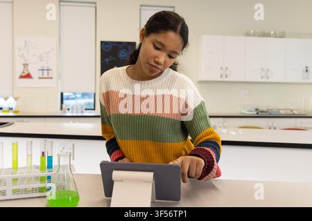 Mädchen in gestreiftem Pullover, das ein Tablet auf einem Ständer in einem Labor mit Reagenzgläsern und einem grünen Kolben benutzt Stockfoto