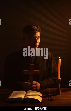 Priester hält Kreuz an hölzernem Tisch mit Heiliger Bibel, Rosenkranz und Kerze vor dunklem Hintergrund Stockfoto