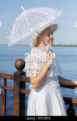 Elegante reife Frau in einem Vintage-weißem Spitzenkleid, die einen Sonnenschirm an einem Flussufer hält. Stockfoto