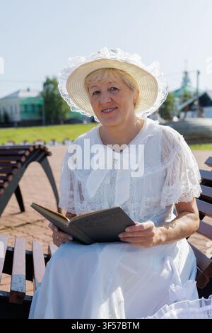 Lächelnde reife Frau in einem Vintage-weißen Spitzenkleid und Hut, die ein altes Buch an einem sonnigen Ufer liest Stockfoto