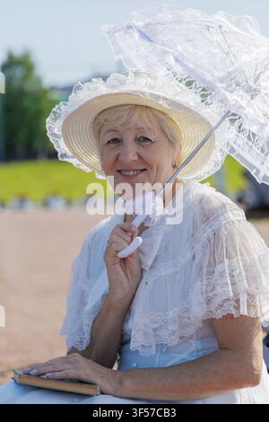 Lächelnde reife Frau in einem Vintage-weißen Spitzenkleid, die auf einer modernen Holzbank mit einem Buch und einem Sonnenschirm sitzt. Stockfoto