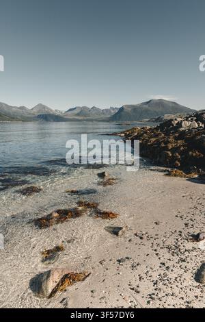 Blick auf die klaren Küstengewässer und die felsige Küstenlinie mit fernen Bergen unter einem klaren Himmel in Sommaroy, Troms, Norwegen. Stockfoto