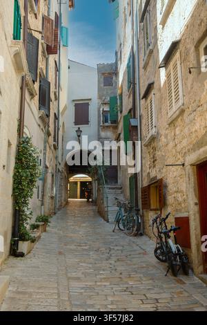 Historische Kopfsteinpflastergasse mit Steinmauern, grünen Fensterläden, gewölbtem Durchgang und geparkten Fahrrädern in Rovinj, Istrien, Kroatien. Stockfoto