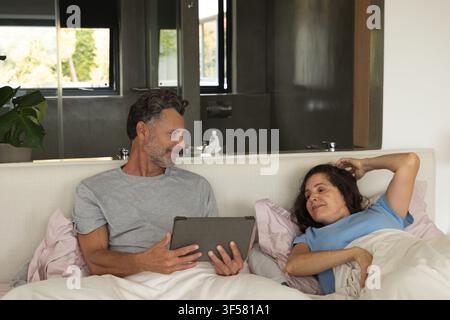 Paar liegt auf dem Bett im Hauptschlafzimmer, Mann hält ein Tablet, Frau liegt unter der Bettdecke Stockfoto