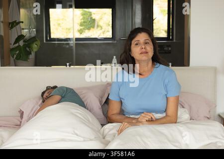 Reife Frau sitzt rechts vom Bett neben ihrem schlafenden Partner zu Hause in einem hellblauen T-Shirt Stockfoto