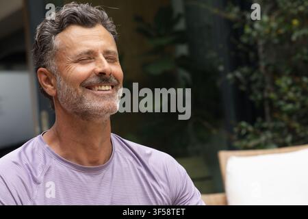 Mittelalter Mann lächelt mit geschlossenen Augen, sitzt auf einem Terrassenstuhl und trägt ein lila Sporthemd Stockfoto