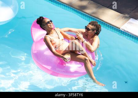 Afroamerikanisches Paar liegt auf einem rosa Floß am Pool, trägt Badeanzüge und hält Plastikbecher Stockfoto