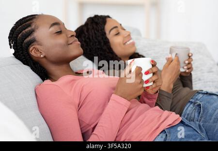 Entspannte afrikanische Damen, die Tee trinken, während sie sich auf der Couch ausruhen Stockfoto