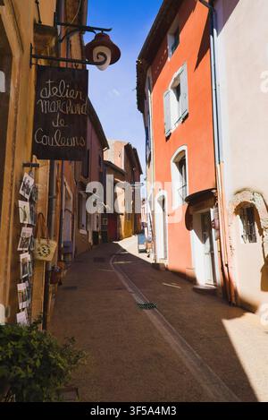 Bezaubernde enge Gasse mit farbenfrohen ockerfarbenen Fassadengebäuden und Ladenschildern im Hügeldorf Roussillon, Provence, Frankreich. Stockfoto